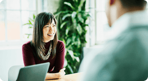 Una mujer sonríe mientras está sentada frente a un colega, en una oficina bien iluminada