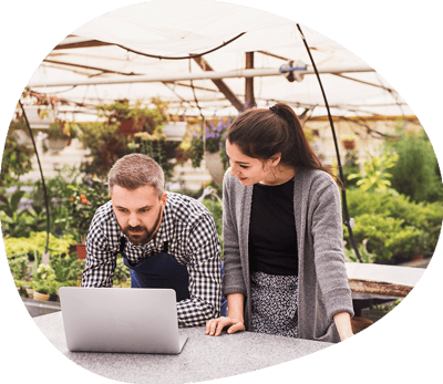 A man and a woman standing in a greenhouse looking at a laptop