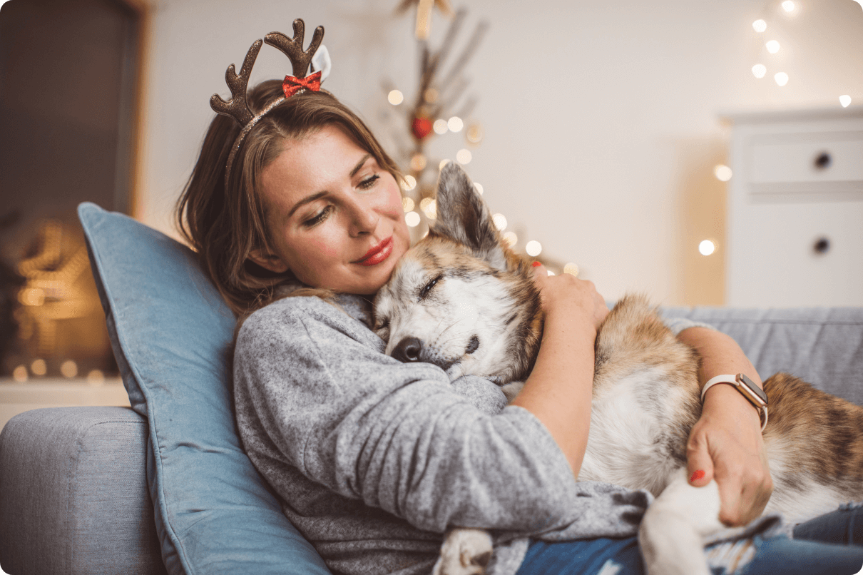 A young woman smiles and is happy as she reflects on the new year with her dog on the couch, holiday lights surround her