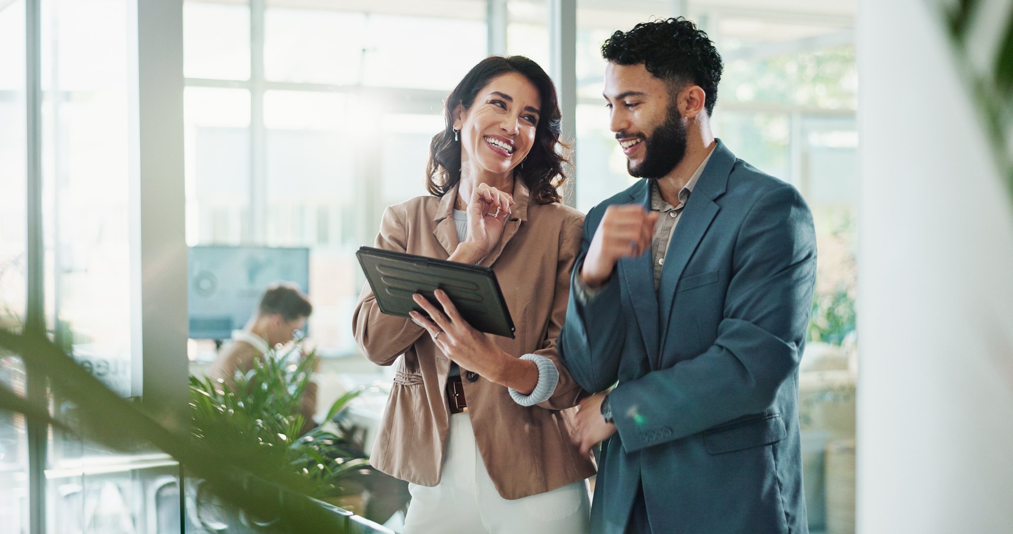 Two professionals stand in a bright, modern office, reviewing information on a tablet together while discussing it, with desks, plants, and coworkers visible in the background.