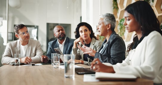 A group of professionals sit around a conference table in a modern office, engaged in discussion with notebooks, documents, and glasses of water in front of them, suggesting a collaborative meeting or planning session.