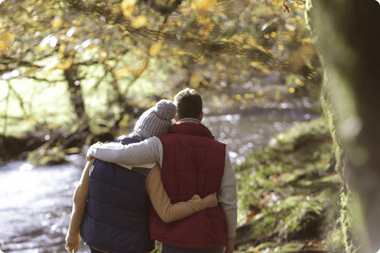 Couple dressed warmly embrace while standing in front of a riverbank. 