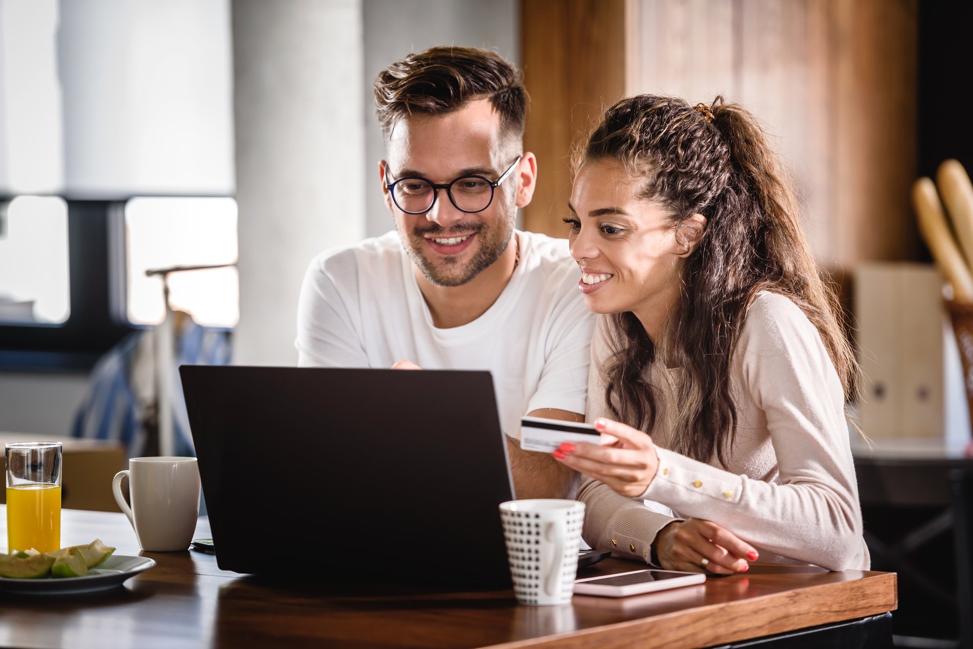 Two people sit at a wooden table in a bright home setting, looking at a laptop while one person holds a benefits accounts card. On the table are a coffee mug, a glass of juice, a plate with food, and a smartphone, suggesting an online purchase or account task.