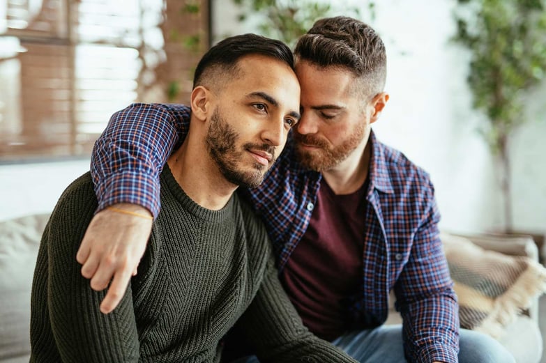 Two sad-looking men sitting on a couch, one man with his arm around the other.