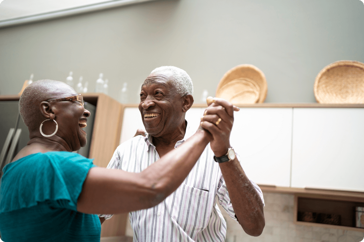 A happy elderly couple dance while holding hands in their kitchen. 