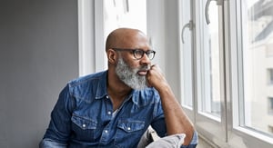 A bald person with a beard, wearing glasses and a denim shirt, gazing thoughtfully out of a window.