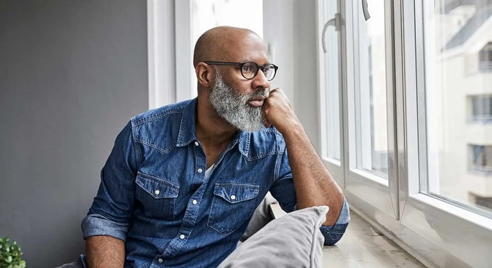 Une personne chauve avec une barbe, portant des lunettes et une chemise en jean, regardant pensivement par la fenêtre.