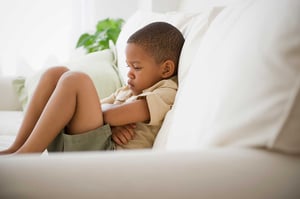 A frustrated-looking little boy sitting on a couch with his arms wrapped around his legs.