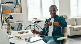 A smiling man in a teal shirt sitting at a desk in a bright, modern home office with a bicycle in the background