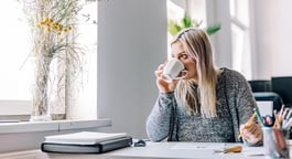A woman with blonde hair sitting at a desk, taking a sip from a white coffee cup.