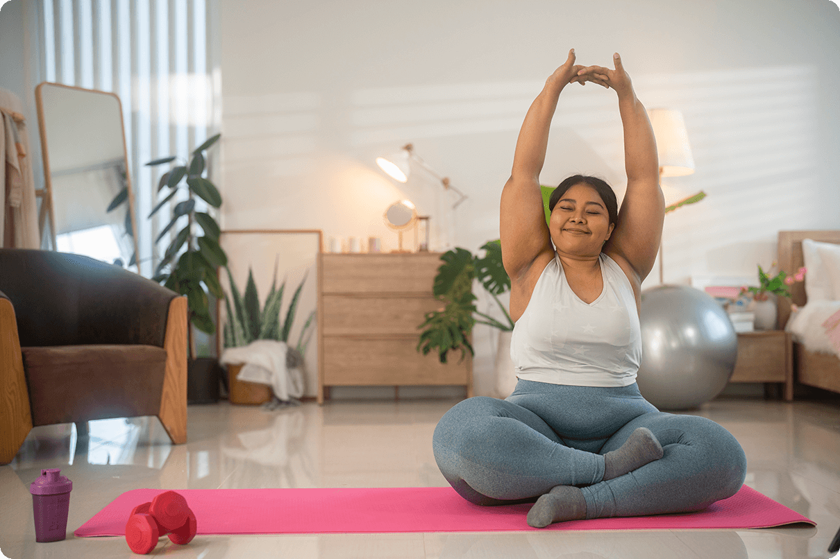 A smiling young lady sits cross-legged on the floor stretching her arms in the air. 