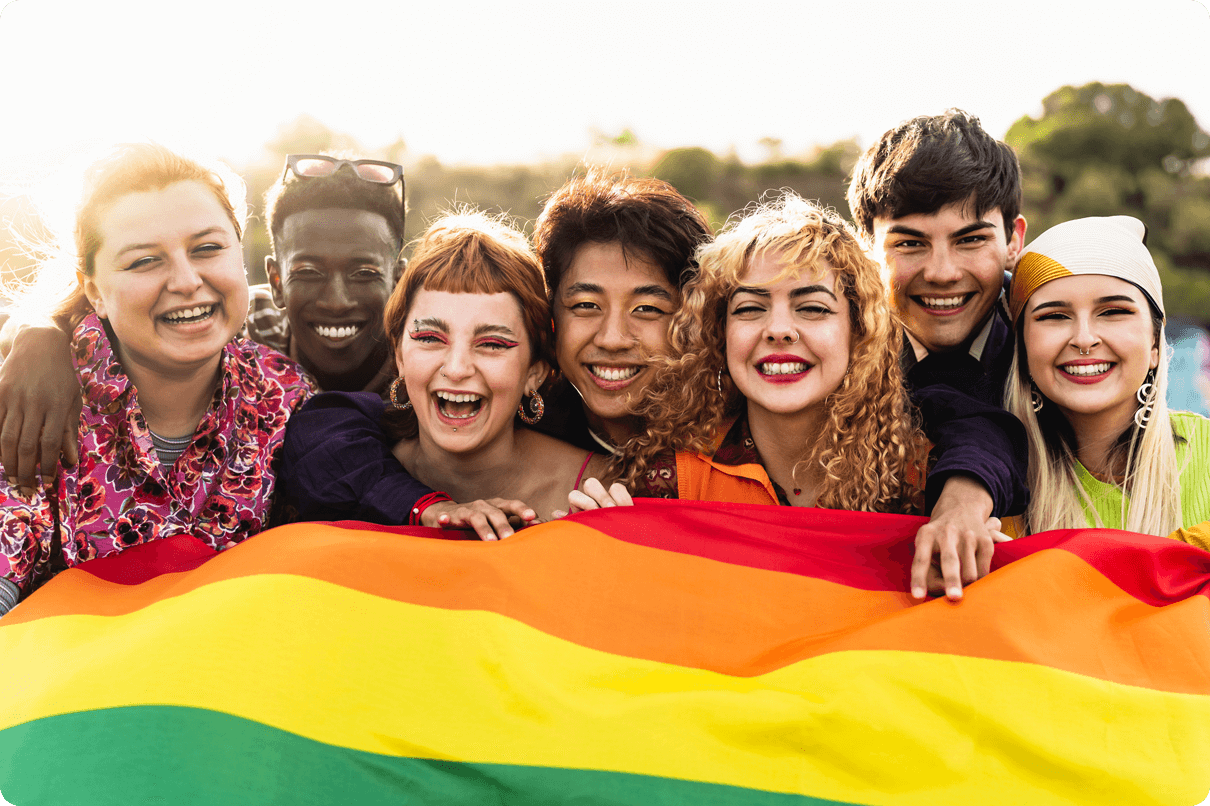 A group of multicultural smiling young people stand in front of a colourful flag. 