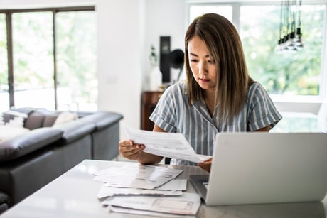 Woman sitting at a kitchen counter looking at a stack of invoices with her laptop open.