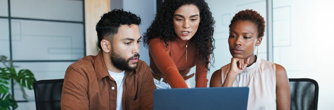  Tres profesionales colaborando en una oficina moderna, dos mujeres y un hombre reunidos alrededor de una laptop, revisando contenido en la pantalla juntos en un ambiente de trabajo colaborativo