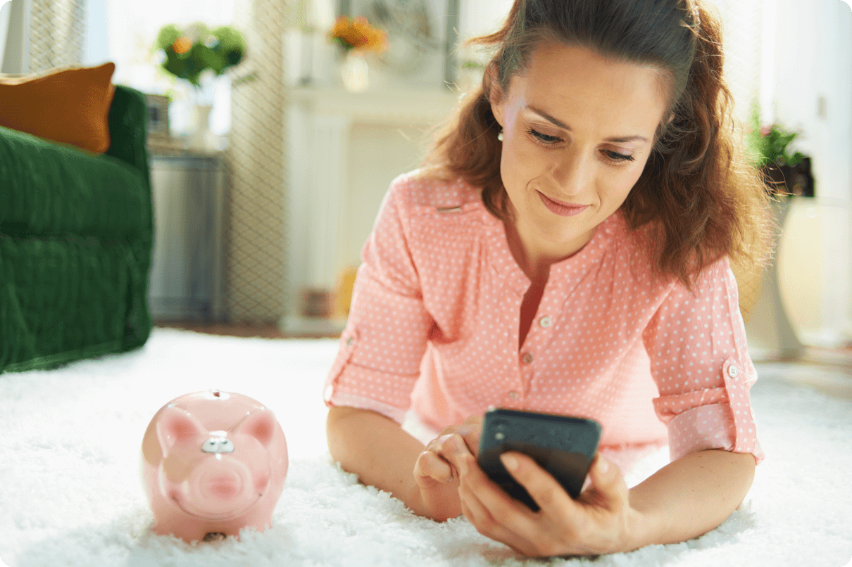 Lady lying on the floor looking at her phone with a pink piggy bank sitting next to her. 