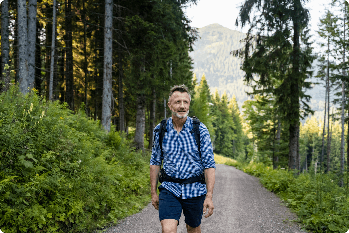 An older man wearing a backpack walks on a trail in the forest. 