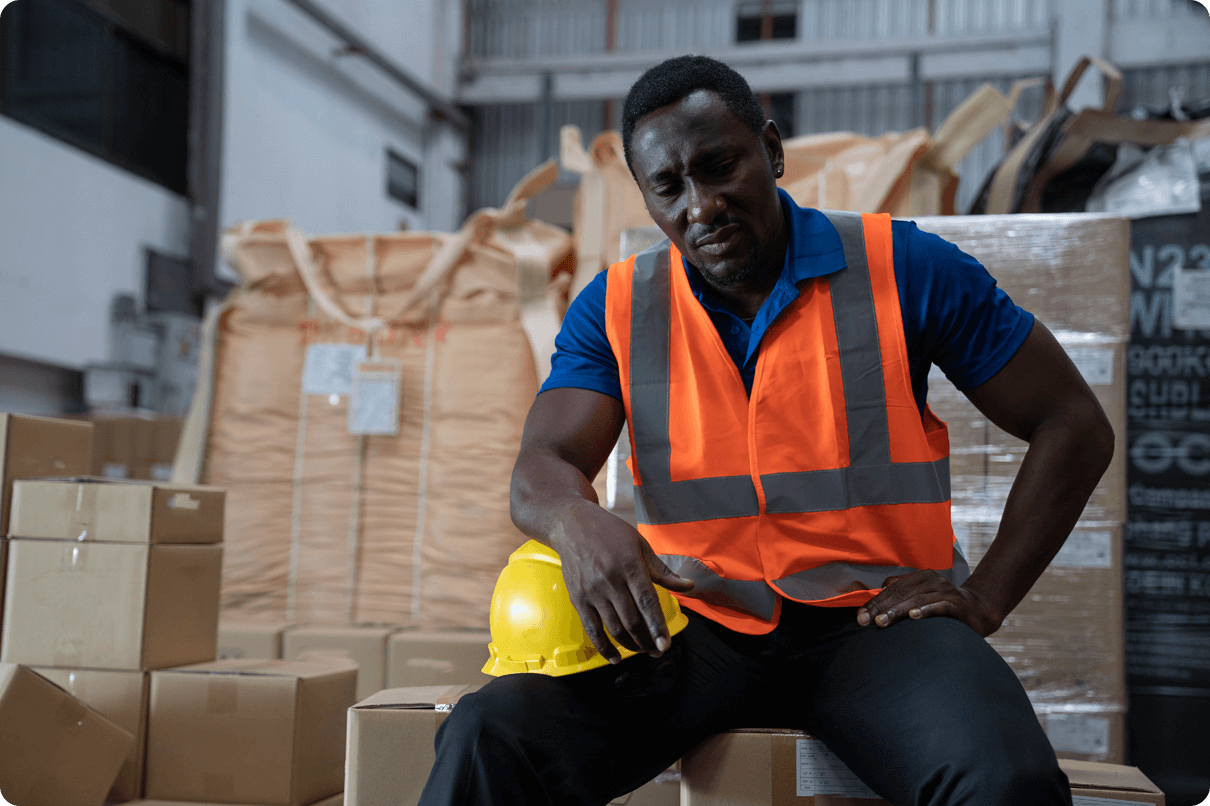 Sad-looking man wearing a work uniform looks down while sitting on a box in a warehouse.