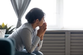 Una mujer de aspecto preocupado mira por la ventana con el rostro entre las manos.