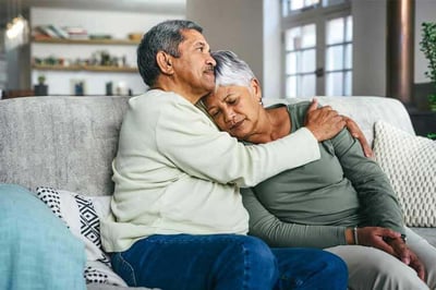 A sombre-looking man and woman embracing while sitting on a couch.