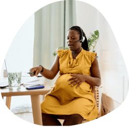 A pregnant woman in a yellow dress sitting at a table, touching her belly with one hand and holding a pen with the other hand over a notebook,