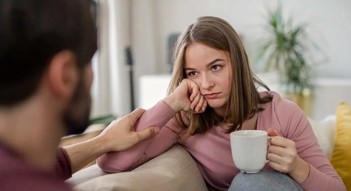 A person sitting on a couch with a person holding a cup.