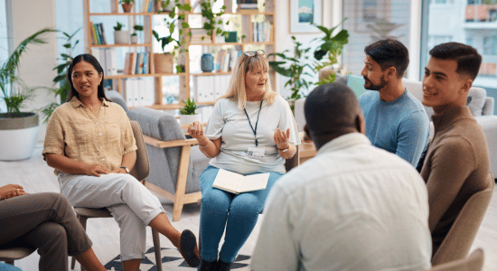 Diverse group of adults seated in a circle, engaged in a group discussion in a bright, plant-filled room.