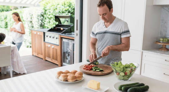 Man slicing cucumbers and tomatoes on a cutting board in a bright kitchen while another person prepares food outside near a grill on a patio.
