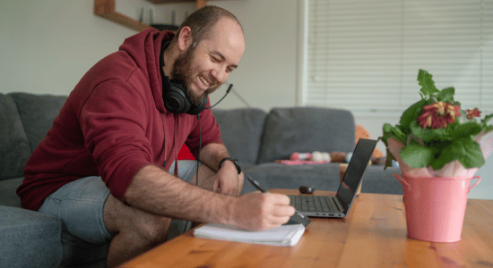 Smiling man in a red hoodie with headphones around his neck writes in a notebook while using a laptop at a coffee table.