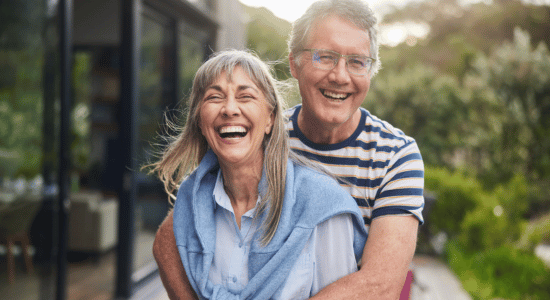An older woman and man smiling and laughing while embracing in a garden setting outside a home