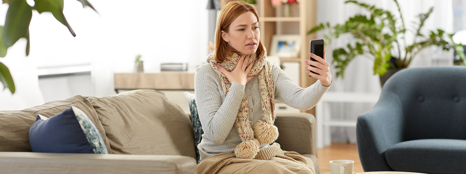Woman with reddish-brown hair sitting on a beige couch in a living room, touching her throat and looking at her smartphone. She's wearing a light gray cardigan and a patterned scarf. The room has plants and bookshelves in the background, with a cup visible on a nearby surface.