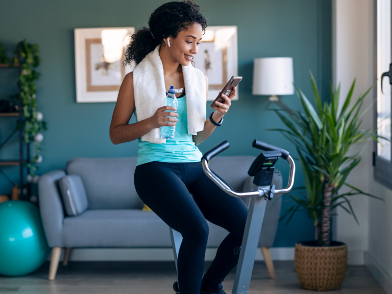 Young fitness girl using mobile phone while training on exercise bike at home