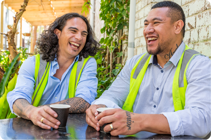 Two construction workers sitting at a table drinking coffee and smiling